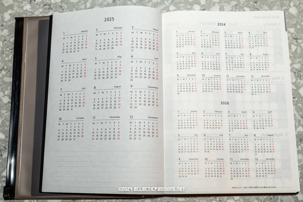 An open planner on a gray-speckled surface displays calendar years 2024, 2025, and 2026. The left page shows 2025, while the right page has 2024 and 2026. The planner is ring-bound and has a glossy cover.