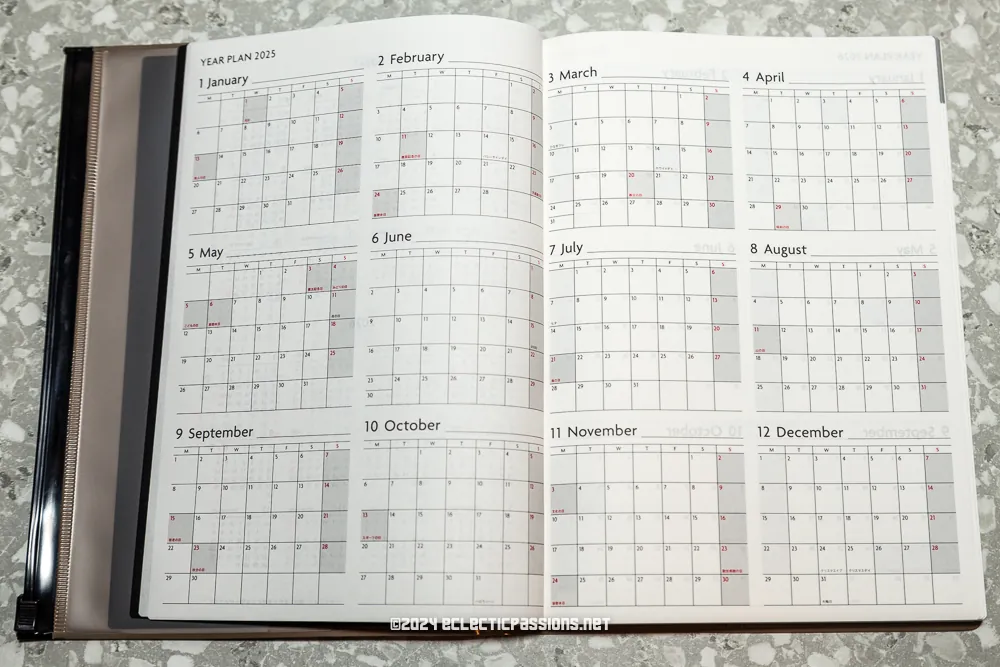 A planner lies open to a year plan for 2025, showing monthly calendars from January to December. Each month is neatly arranged with dates in a grid format. The planner features a grey speckled background.