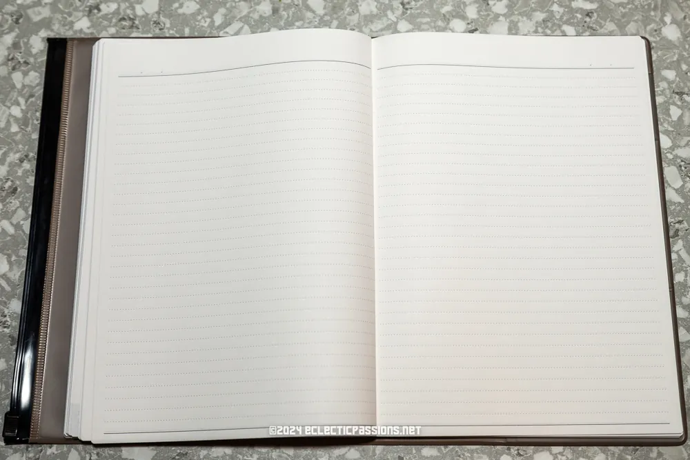 An open notebook with lined pages lies flat on a speckled gray surface. The binding is on the left, and the notebook appears unused. At the bottom center of the open pages is a faint watermark with text.