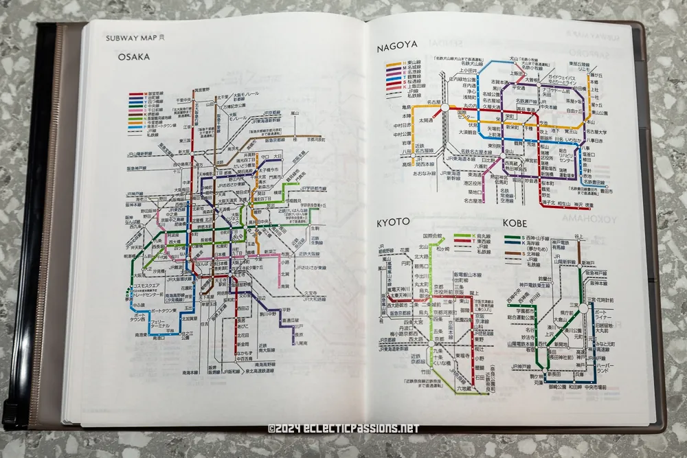 An open book displaying detailed subway maps for Osaka, Nagoya, Kyoto, Kobe, and Amagasaki. The maps include various colored lines and station names in both Japanese and English, set against a speckled gray background.