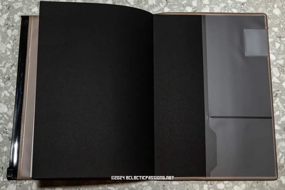 An open, empty black album with transparent plastic pockets on a speckled surface. The album's spine is visible on the left, and a partially obscured web address is seen at the bottom.