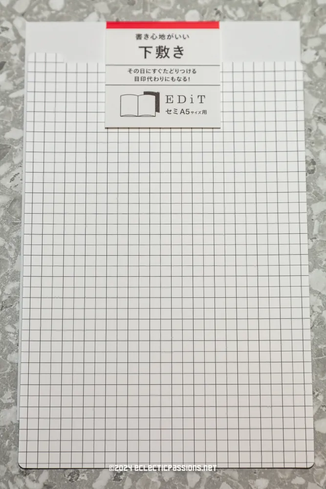 A grid-patterned notebook with a label featuring Japanese text and the word "EDiT" in English. The notebook is placed on a speckled surface.