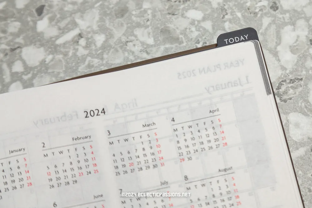 Close-up of a planner open to yearly calendars for 2024 and 2025 on a speckled gray surface. A tab labeled "Today" is visible in the upper right corner. The planner shows months with days of the week marked.