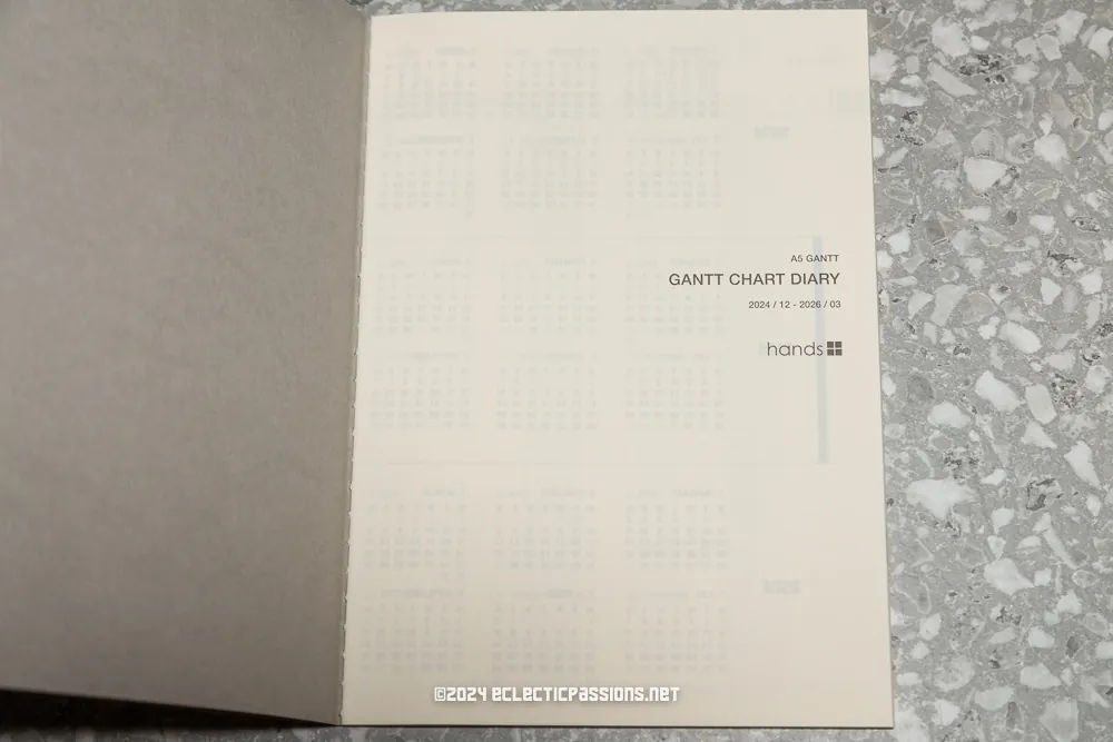Open notebook displaying a Gantt chart diary title page with the text "A6 Gantt Gantt Chart Diary 2021/12 - 2022/03" and "hands". The background shows a terrazzo-patterned surface.