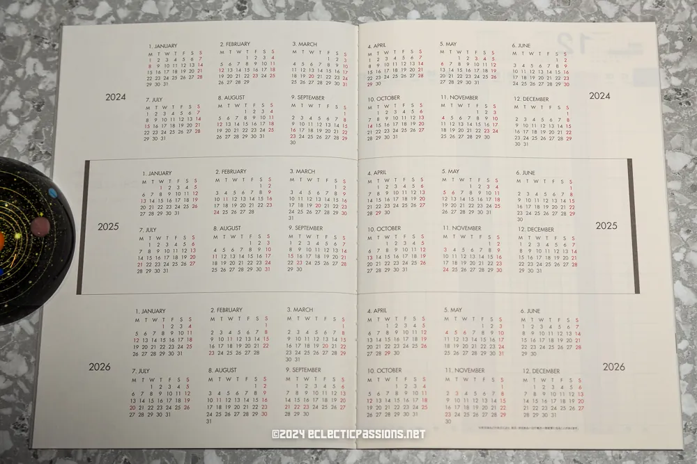 A 2024-2026 yearly calendar spread open on a marble surface. The calendar displays months from January 2024 to December 2026 in a compact grid format.