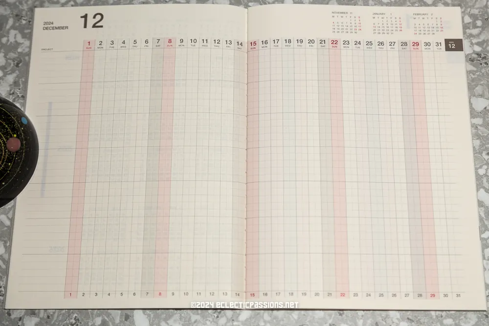 An open planner displaying a monthly calendar view for December 2023. The layout includes numbered days in columns, with weekends in red. The planner rests on a textured gray surface, and a round object is partially visible on the left.