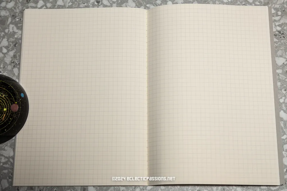 An open notebook with grid paper is centered on a speckled surface. A black mug with a celestial design sits to the left of the notebook.