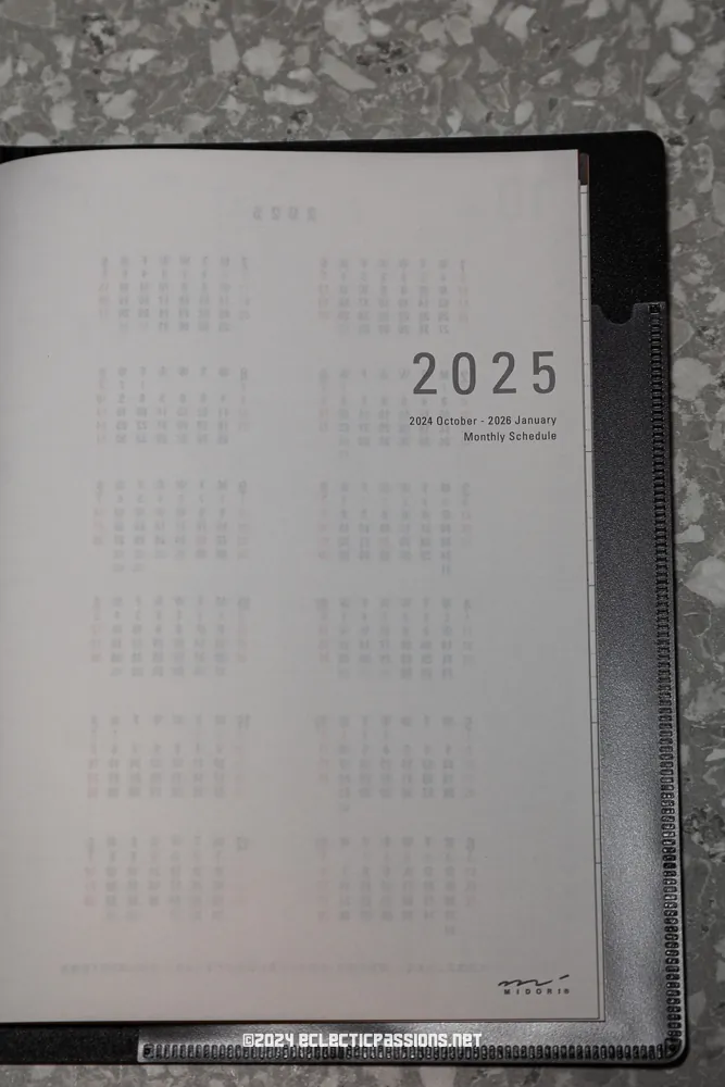 A close-up of an open planner on a grey patterned surface, displaying a page with "2025" and the text "2024 October - 2026 January Monthly Schedule." Calendar months are printed in small font.