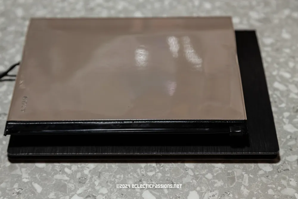 A stack of two tablets or electronic devices lies on a speckled gray surface. The top device has a shiny, reflective surface with a zipper partially visible on the edge. The bottom device has a textured dark finish.