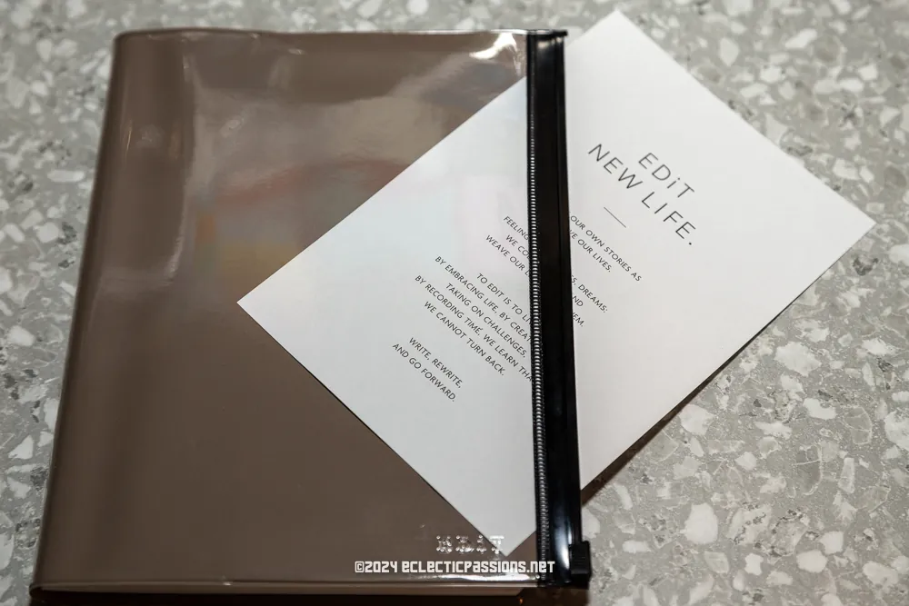 A transparent plastic folder partially reveals a white card print. The card reads "Edit New Life" and lists topics such as choosing tools and exploring spaces. The background is a speckled gray surface.